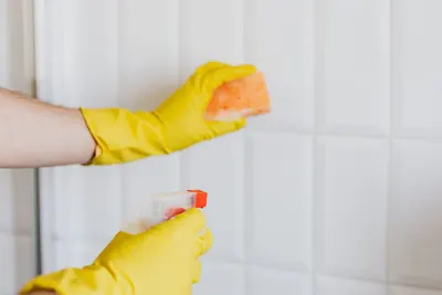 Yellow gloved hands holding a sponge and spray bottle at white wall tile